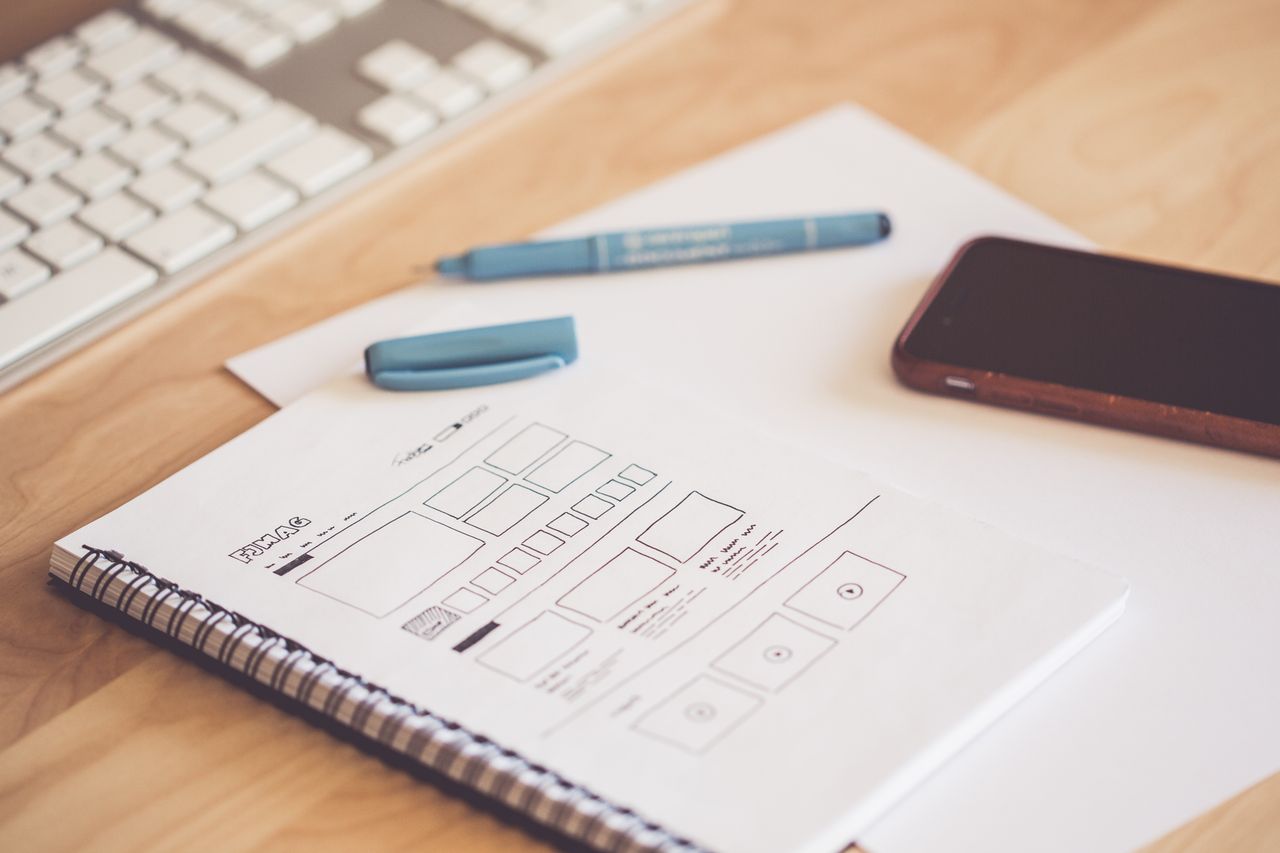 about-us-02 A neat workspace featuring a sketchpad with wireframes, a smartphone, and a keyboard on a wooden desk.