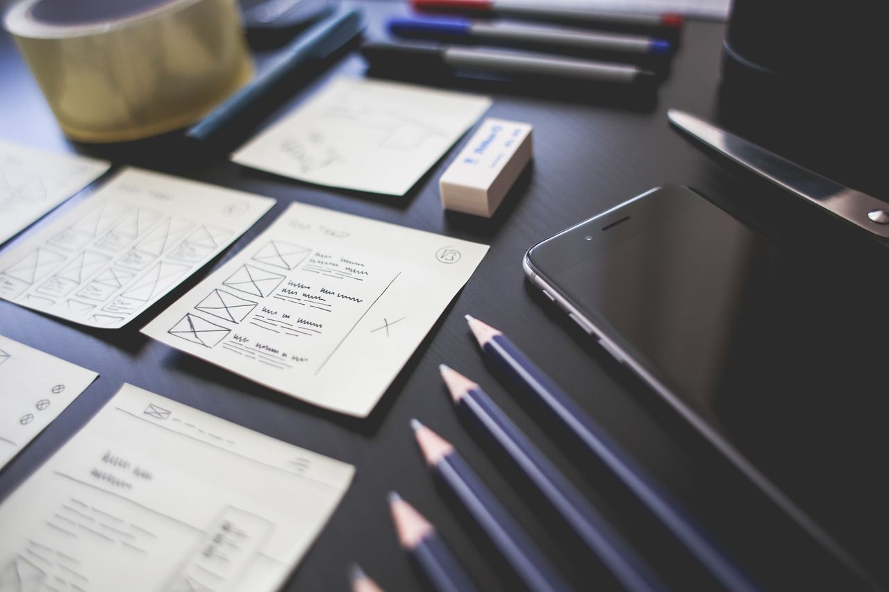 about-us-01 A creative workspace featuring UI design plans, pencils, a smartphone, and other office tools.